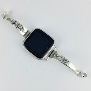 Op de afbeelding: Zilveren horlogeband met een sierlijk ontwerp. Het bandje is bevestigd aan een vierkante smartwatch met een zwart scherm en het woord "Fitbit" gedrukt aan de onderkant.