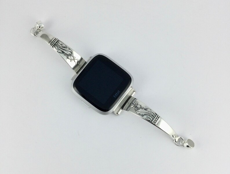 Op de afbeelding: Zilveren horlogeband met een sierlijk ontwerp. Het bandje is bevestigd aan een vierkante smartwatch met een zwart scherm en het woord "Fitbit" gedrukt aan de onderkant.