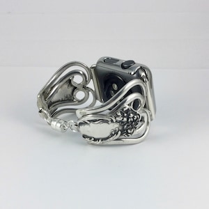 May include: A silver Apple Watch band crafted from repurposed silverware. The band features ornate floral and scroll designs, with a clasp closure. The watch face is a dark gray color, and the band is designed to fit the watch securely.