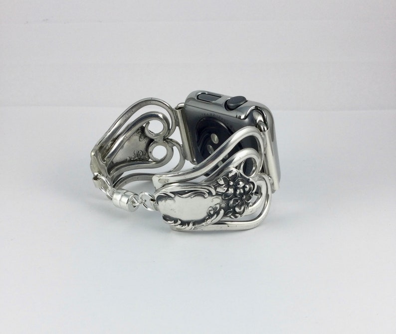 May include: A silver Apple Watch band crafted from repurposed silverware. The band features ornate floral and scroll designs, with a clasp closure. The watch face is a dark gray color, and the band is designed to fit the watch securely.