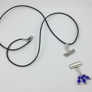 May include: A black cord necklace with a silver-colored clasp and pendant. The pendant has a silver-colored metal clasp with a cluster of blue crystal beads. The necklace is displayed on a white background.