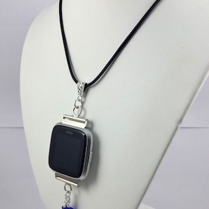 May include: A silver Fitbit watch pendant necklace with a black band. The watch face is black. The pendant has a silver chain with blue crystal beads hanging from the bottom. The necklace is displayed on a white mannequin.