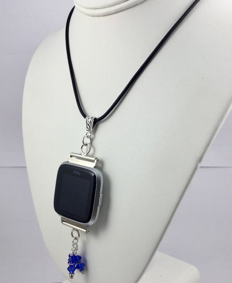May include: A silver Fitbit watch pendant necklace with a black band. The watch face is black. The pendant has a silver chain with blue crystal beads hanging from the bottom. The necklace is displayed on a white mannequin.