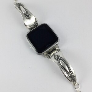 May include: Silver watch band made from a vintage spoon with a black Fitbit watch face. The band has a floral design and the watch face says "Fitbit".