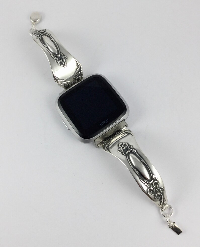 May include: Silver watch band made from a vintage spoon with a black Fitbit watch face. The band has a floral design and the watch face says "Fitbit".
