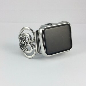 May include: Silver-toned Apple Watch band with an ornate floral design on the left side. The band is connected to a silver watch face with a black screen. The watch and band are on a white background.