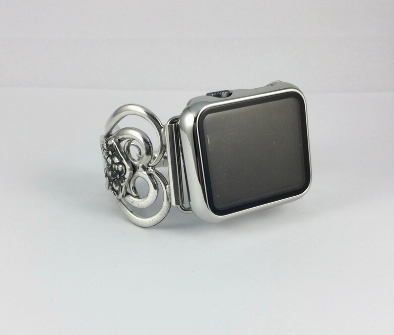 May include: Silver-toned Apple Watch band with an ornate floral design on the left side. The band is connected to a silver watch face with a black screen. The watch and band are on a white background.