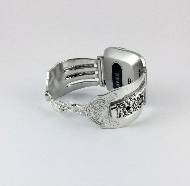 May include: Silver smartwatch band made from a repurposed fork with an ornate design. The band is attached to a silver smartwatch with a black screen.