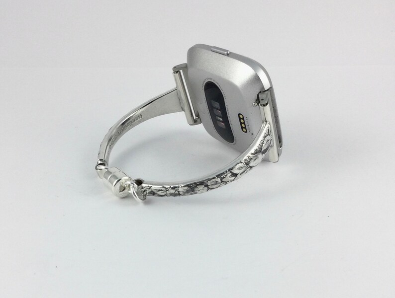 May include: A silver bracelet with a floral design that holds a silver smartwatch.