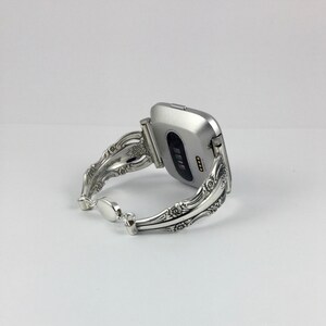 May include: Silver watch band made from a repurposed silverware piece. The band is ornate and features a floral design. The watch face is silver and square shaped.
