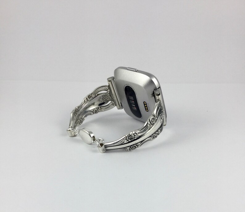 May include: Silver watch band made from a repurposed silverware piece. The band is ornate and features a floral design. The watch face is silver and square shaped.