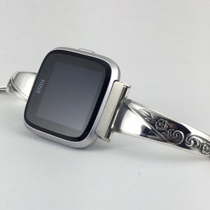 May include: A silver Fitbit smartwatch with a floral-patterned silver band. The band is made from a repurposed spoon.