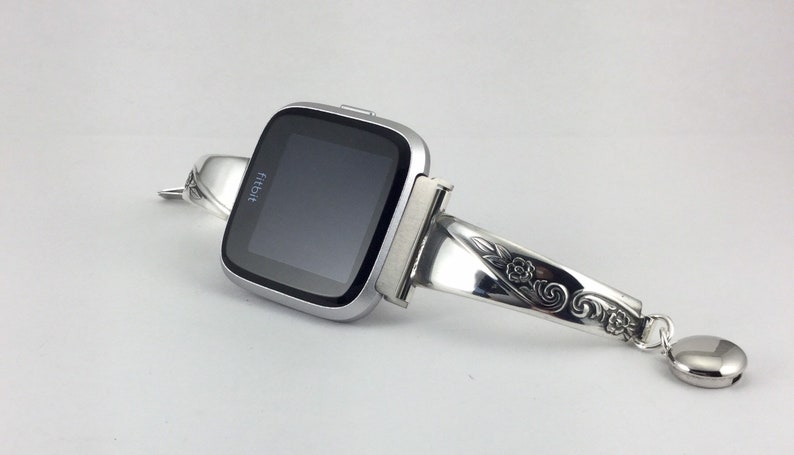 May include: A silver Fitbit smartwatch with a floral-patterned silver band. The band is made from a repurposed spoon.