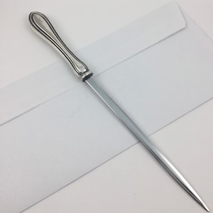 May include: A silver letter opener with a decorative handle and a sharp blade. The letter opener is lying on a white envelope.