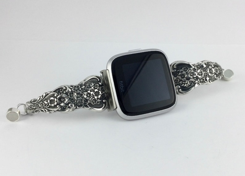 May include: A silver Fitbit smartwatch with a decorative floral band. The band features intricate flower and leaf designs. The watch has a square face with a black screen and the Fitbit logo.
