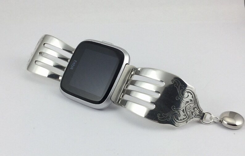 Pu&ograve; includere: Un orologio intelligente Fitbit argentato con un braccialetto personalizzato a forma di forchetta. Il braccialetto presenta un design decorativo e un ciondolo argentato.
