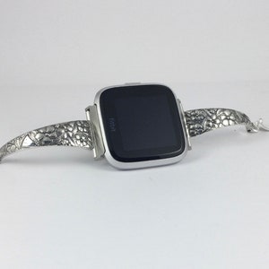 May include: A silver Fitbit smartwatch with a textured silver band. The band has a floral design.