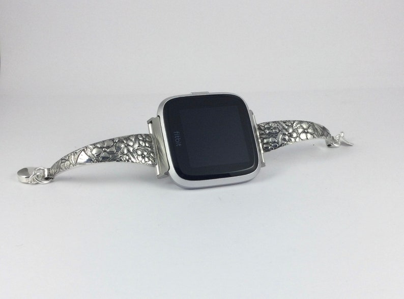 May include: A silver Fitbit smartwatch with a textured silver band. The band has a floral design.