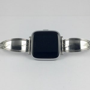 May include: A silver smartwatch with a black screen is attached to a silver spoon band. The band is made from two spoons that have been bent into a loop and attached to the watch.