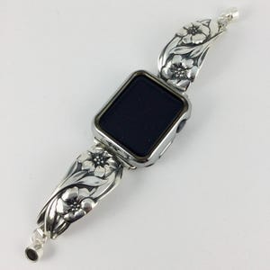 May include: A silver watch strap featuring a floral pattern, attached to a smartwatch with a black screen. The floral design includes detailed leaves and flowers. The strap is made of a shiny metal.