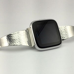 K&ouml;nnte beinhalten: Eine silberne Fitbit Smartwatch mit einem strukturierten silbernen Armband. Das Armband ist gebogen und aus geh&auml;mmertem Metall.