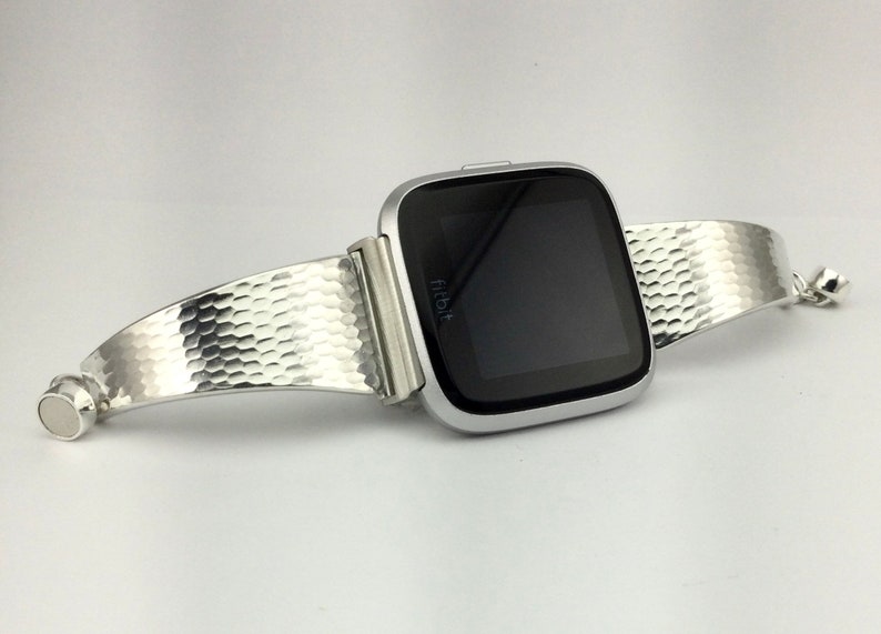 K&ouml;nnte beinhalten: Eine silberne Fitbit Smartwatch mit einem strukturierten silbernen Armband. Das Armband ist gebogen und aus geh&auml;mmertem Metall.