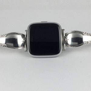 May include: A silver smartwatch with a black screen is attached to a silver bracelet made from two spoons. The bracelet has a decorative design and a clasp.