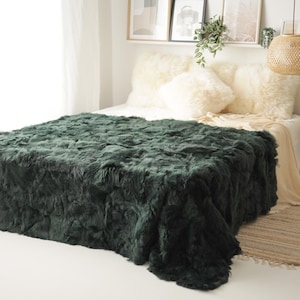 May include: A plush, dark green faux fur throw blanket on a bed with white pillows. The blanket is soft and fluffy, and it would be perfect for adding a touch of luxury to any bedroom.