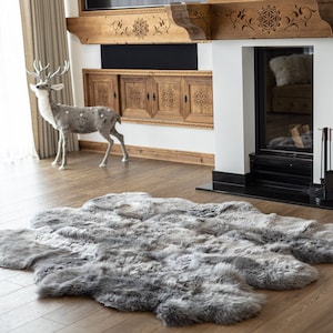 Puede incluir: Una alfombra grande de piel de oveja gris con textura peluda, colocada sobre un suelo de madera en un salón. La alfombra está delante de una chimenea con azulejos negros y una repisa de madera.
