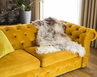 Può includere: Un divano giallo senape trapuntato con una coperta in pelle di pecora e due cuscini abbinati. Il divano ha borchie argentate sui braccioli. È appoggiato su un pavimento in legno. Il divano misura circa 200 cm di lunghezza.