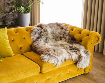 Può includere: Un divano in velluto giallo con una coperta in pelle di pecora drappeggiata sopra. Il divano ha bottoni capitonné e bordi chiodati. Ci sono due cuscini gialli sul divano.