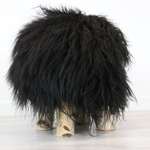 May include: Black sheepskin ottoman with four birch wood legs. The ottoman is round and fluffy.