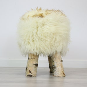 May include: A white sheepskin ottoman with four birch wood legs. The ottoman is round and fluffy.