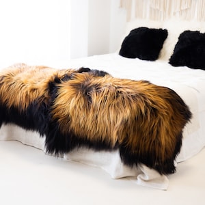 May include: Two sheepskin rugs in black and brown tones, placed on a white bed with black and white throw pillows. The rugs are made of soft, fluffy fur.