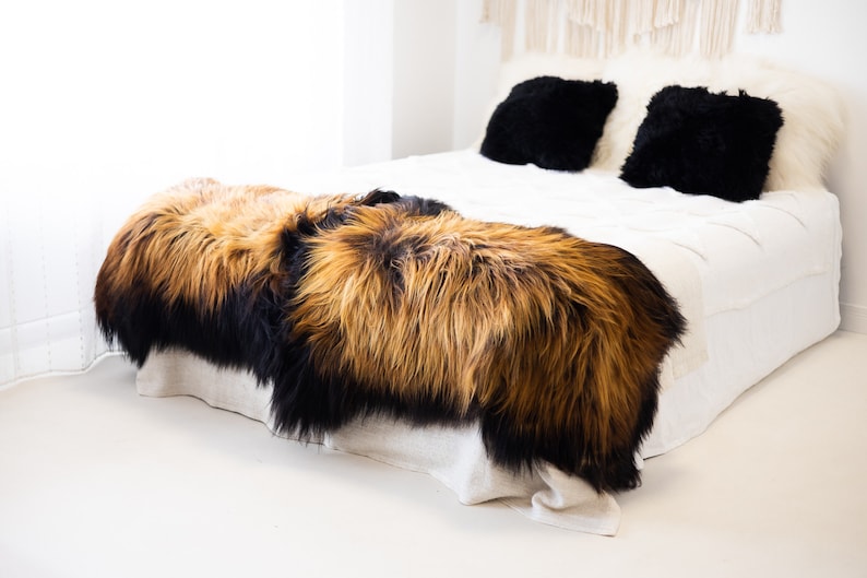 May include: Two sheepskin rugs in black and brown tones, placed on a white bed with black and white throw pillows. The rugs are made of soft, fluffy fur.