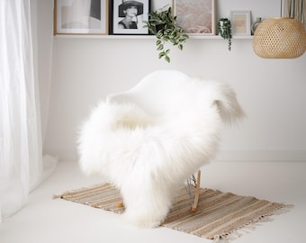 May include: A white sheepskin rug draped over a white chair in a minimalist room with a woven pendant light hanging from the ceiling.