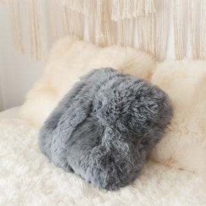 May include: A selection of soft, textured cushions in grey and cream hues. The cushions are placed on a fluffy, white, shaggy rug, creating a comfortable and welcoming feel. Decorative elements are visible in the background.