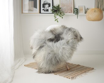 May include: A fluffy, gray and white faux fur throw draped over a white chair. The chair sits on a small, rectangular, woven rug. The background includes framed artwork and a woven pendant light, creating a cozy, neutral-toned interior design.