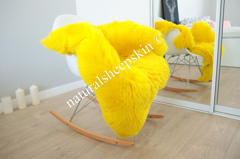 May include: A bright yellow sheepskin rug draped over a white rocking chair. The rug is soft and fluffy, and the chair is made of metal and wood. The text "naturalsheepskin" is visible on the rug.