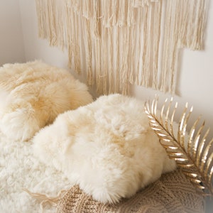 May include: Two fluffy white faux fur throw pillows on a white faux fur bedspread with a macrame wall hanging in the background.