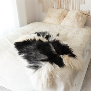 Peut inclure: Un lit blanc avec un tapis en peau de mouton noir et blanc sur le dessus. Le lit a des oreillers blancs et moelleux et une couverture blanche. Le lit est dans une chambre avec des murs blancs et une fenêtre en arrière-plan.