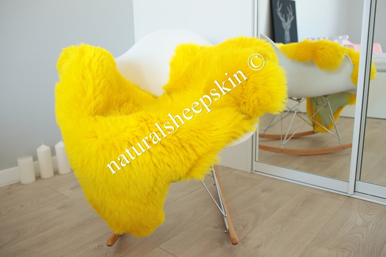 May include: A bright yellow sheepskin rug draped over a white rocking chair. The rug is soft and fluffy, and the chair is made of metal and wood. The text "naturalsheepskin &copy;" is visible on the rug.