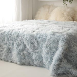 Op de afbeelding: Een lichtblauwe imitatiebont deken bedekt een bed, met een zachte, donzige textuur. De deken is over het bed gedrapeerd, met witte kussens op de achtergrond. Een gezellig woondecoratie-item.