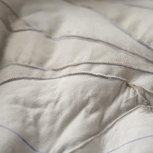 May include: Close-up of a light grey, textured fabric with visible stitching. The fabric has a quilted appearance with curved, horizontal lines of stitching. Blue lines are visible between the stitched sections, creating a pattern.