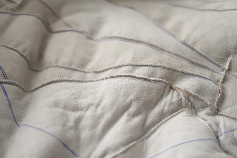 May include: Close-up of a light grey, textured fabric with visible stitching. The fabric has a quilted appearance with curved, horizontal lines of stitching. Blue lines are visible between the stitched sections, creating a pattern.