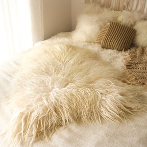 Op de afbeelding: Een gezellige bedopstelling met een crèmekleurige, harige imitatiebont deken. Het bed is versierd met zachte kussens en een gebreide deken in beige en crèmetinten. De scène roept een gevoel van warmte en comfort op.