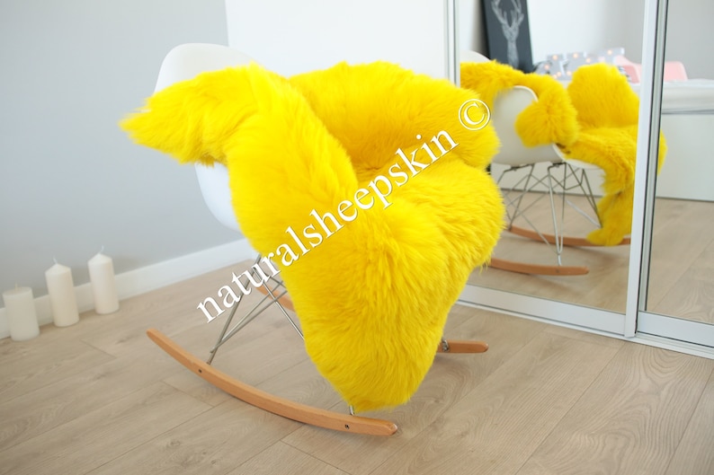 May include: A bright yellow sheepskin rug draped over a white rocking chair. The rug is soft and fluffy, and the chair is made of metal and wood. The text "naturalsheepskin" is visible on the rug.