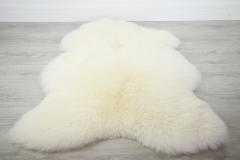 Puede incluir: Una alfombra de piel de oveja blanca con una textura suave y esponjosa. La alfombra est&aacute; extendida sobre un suelo de madera clara.