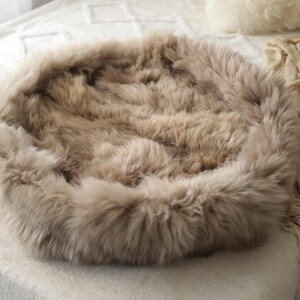 May include: A circular pet bed in a neutral beige hue. The bed features a soft, fluffy texture, with a raised edge and a sunken centre. The bed is designed for comfort and relaxation for small pets.