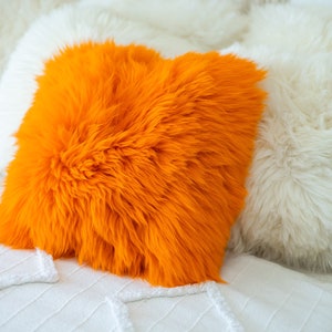 May include: Two fluffy pillows, one orange and one white, on a white bedspread. The orange pillow is in the foreground and the white pillow is behind it.
